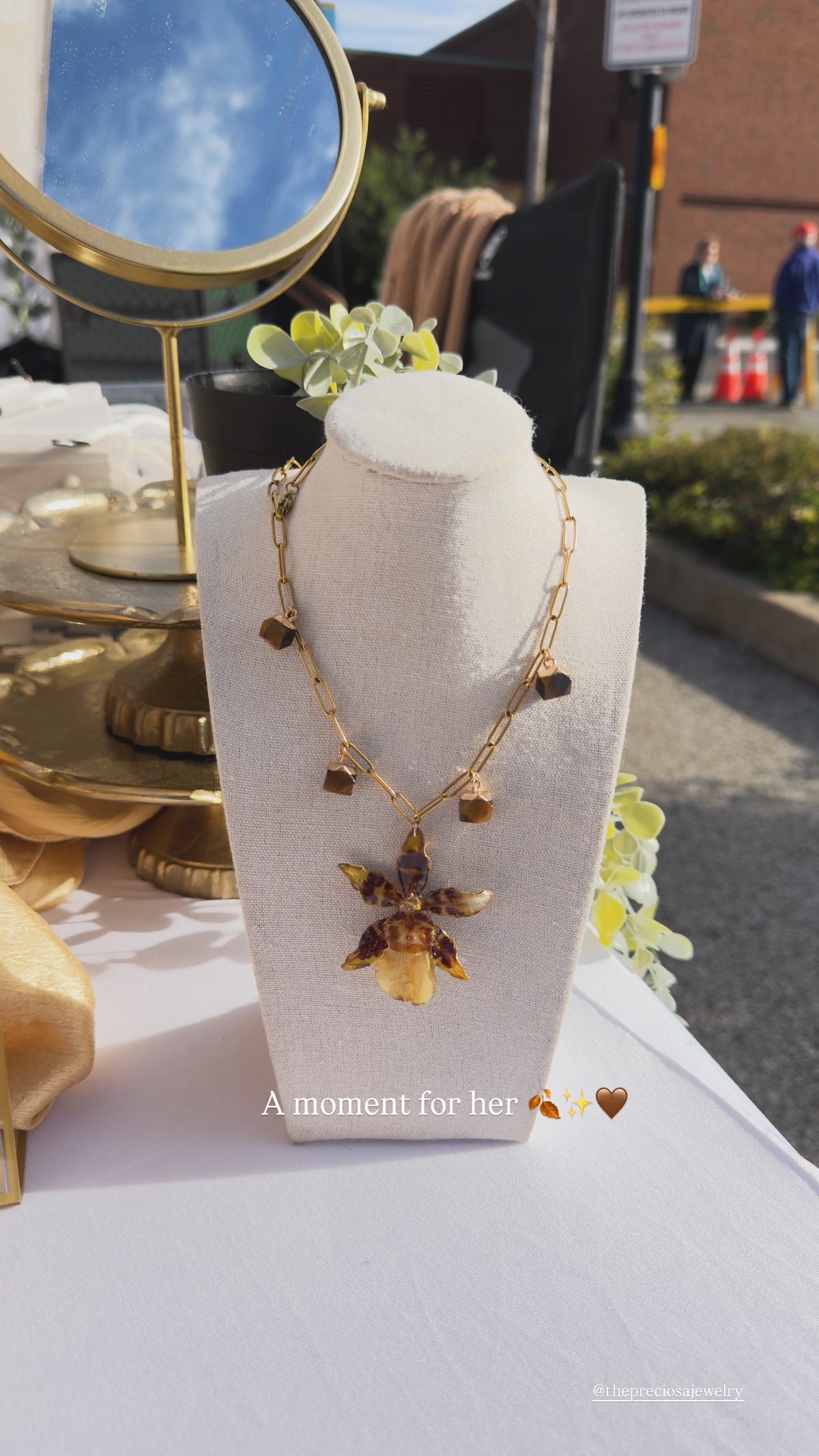 A gold chain necklace with a butterfly pendant displayed on a white fabric mannequin bust. The background shows an outdoor market or cafe setting with a gold mirror and blurred people.