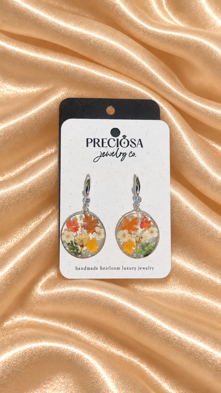 A pair of round dangle earrings displayed on a white card with the brand name 'Preciosa Jewelry Co.' The earrings feature colorful floral and leaf designs inside clear domed circles with silver-toned hooks, set against a draped champagne-colored satin fabric background.